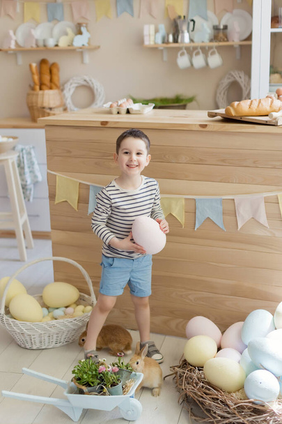 Paskalya süslemeleriyle donatılmış hafif bir mutfakta sevimli bir tavşanı olan beyaz bir çocuk. - Fotoğraf, Görsel