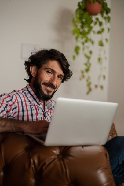 Young business man working at home with laptop on a brown arm chair. Gray notebook for working. Home office concept. High quality photo - Φωτογραφία, εικόνα