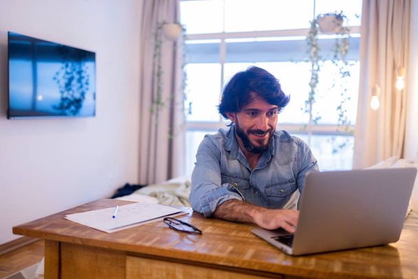 Mladý obchodník pracující doma ve své ložnici s laptopem a papíry na stole. Šedý zápisník pro práci. Koncept domácí kanceláře. Kvalitní fotografie - Fotografie, Obrázek