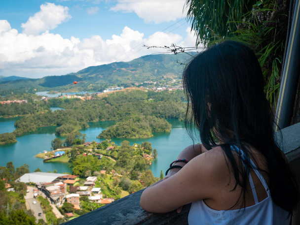 Ciemnowłosa kobieta w białej koszulce spogląda przez balkon z widokiem na Penol - zbiornik wodny Gwatape, Kolumbia - Zdjęcie, obraz