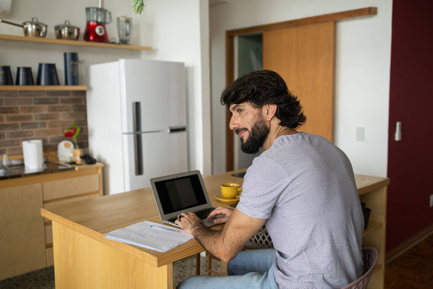 Nuori liikemies työskentelee kotona keittiössään kannettavan tietokoneen ja papereiden kanssa keittiön puupöydällä. Harmaa muistikirja työntekoon. Pääkonttorin konsepti. Laadukas kuva - Valokuva, kuva