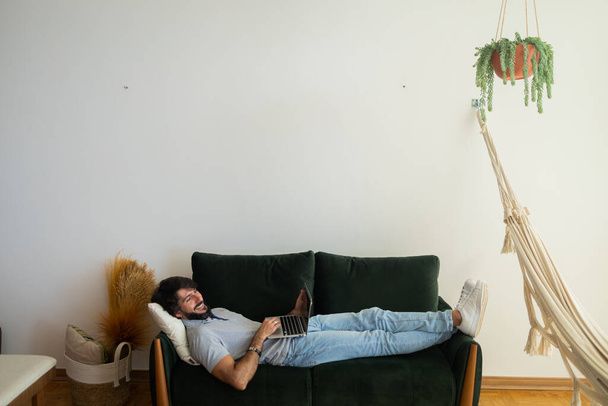 Young business man working at home from his living room couch with laptop on his lap. Man lying down. Gray notebook for working. Green couch. Home office concept. High quality photo - Foto, Bild