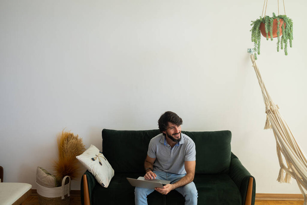 Jeune homme d'affaires travaillant à la maison à partir de son canapé salon avec ordinateur portable sur ses genoux. Carnet gris pour travailler. Concept de bureau à domicile. Photo de haute qualité - Photo, image