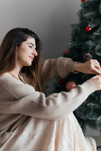 Mladá krásná bruneta v hnědém svetru zdobí vánoční stromek hračkami a je v očekávání dovolené. Šťastný Nový rok, dovolená pozadí - Fotografie, Obrázek