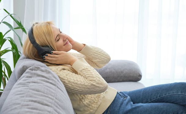 Chica escuchando música con auriculares tumbados en el sofá en casa. - Foto, Imagen