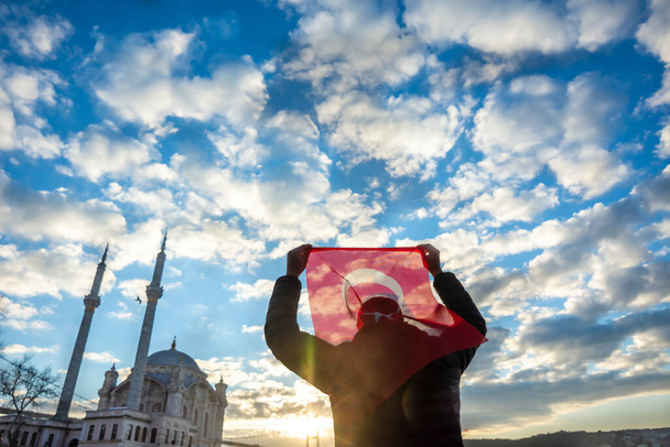 De man heeft de Turkse vlag. Sunrise near Bosphorus bridge (alias: 15 juli Martelaarsbrug Turks: 15 Temmuz Sehitler Koprusu) en de keizerlijke moskee van Sultan Abdulmecid in Ortakoy, Istanbul, Turkije. Patriottisme - Foto, afbeelding
