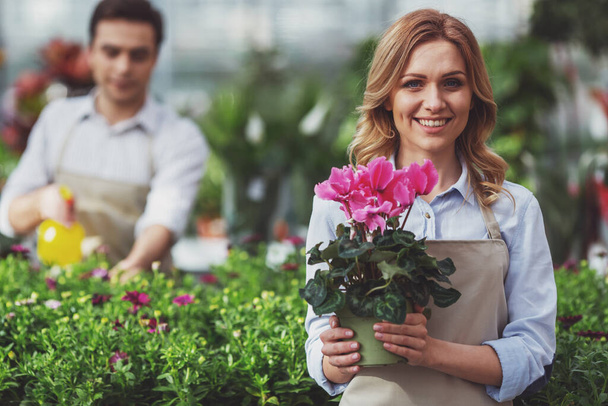 Beautiful young woman is holding a plant and smiling while standing in orangery, in the background man is spraying water on plants - Photo, Image