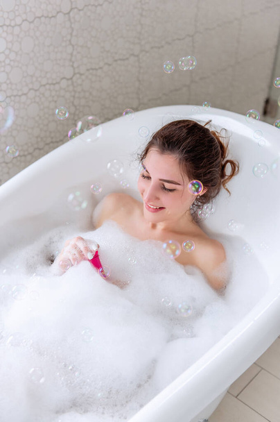 A young woman with hair pinned up in a bun takes a bath with foam, blows, catches soap bubbles. Millennial girl, laughing, having fun. Health hygiene, spa concept, body relaxation. Luxury holiday - Foto, afbeelding