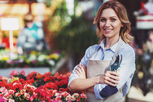 schöne junge Frau in Schürze hält eine Gartenschere, blickt in die Kamera und lächelt, während sie in der Orangerie steht - Foto, Bild