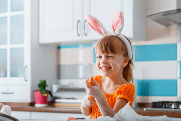 Emotional portrait of cheerful little girl dressed as bunny with carrot for Easter while cooking food in kitchen at home. Girl kid child having fun laughs plays with flour and carrot. - Zdjęcie, obraz