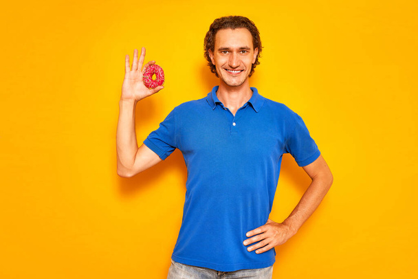 glimlachende man houdt donut in zijn hand in de vorm van een OK-teken. Hij is gekleed in casual kleding, blauwe polo, jeans. geïsoleerd op gele studioachtergrond met ruimte voor tekst. concept - mensen, voedsel, snoep - Foto, afbeelding