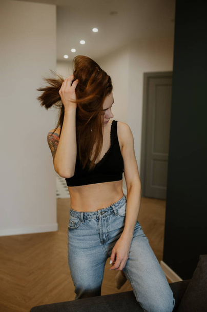 junge schlanke Frau mit roten Haaren, bauchfreiem Oberteil und Jeans in einer stilvollen geräumigen Wohnung scandi modernen Stil. gemütliche Atmosphäre zu Hause. vertikaler, selektiver Fokus - Foto, Bild