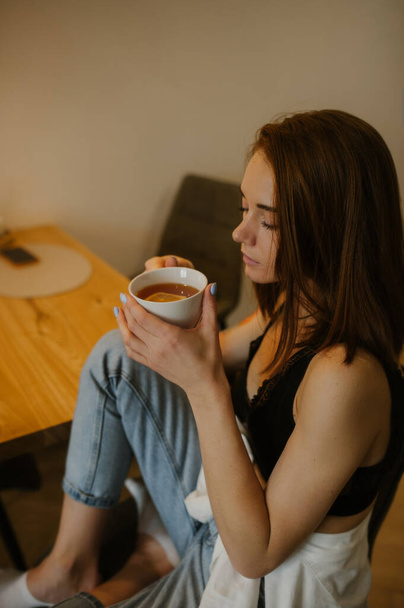 молодая стройная женщина с рыжими волосами сидит за обеденным столом из натурального дерева и пьет чай с лимоном из белой керамической кружки. стильная кухня. Домашняя чайная вечеринка. вертикально, избирательный фокус - Фото, изображение