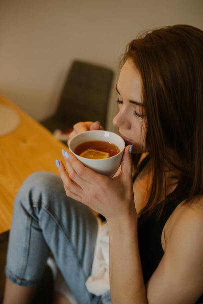 young slender woman with red hair is sitting at natural wood dining table and drinking tea with lemon from white ceramic mug. stylish kitchen. homemade tea party. vertically, selective focus - Foto, Imagen