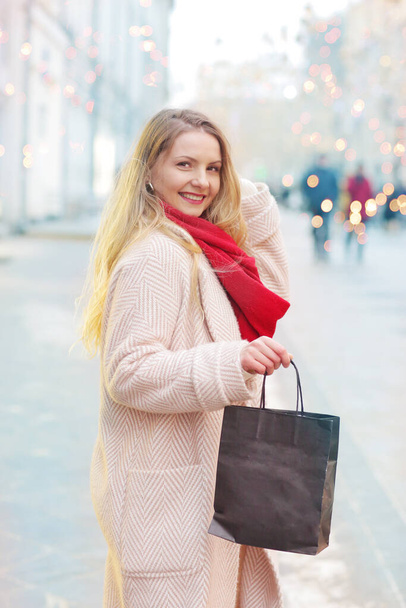 счастливая блондинка в красном шарфе держать торговый мешок с копировальным местом крупным планом фото на рождественском фоне города - Фото, изображение