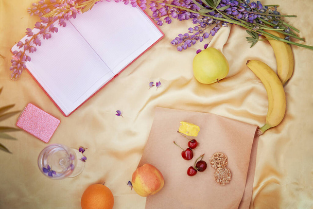 Open notitieboekje mock up met wijnglas, lupine paarse bloemen, appel, bananen, kers, halva, koekjes op gouden achtergrond. Zomer picknick. Plaats voor tekst. Dicht bij de natuur, concept van zelfontdekking - Foto, afbeelding