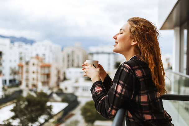 Mladá brunetka oblečená ve spacáckém oblečení si vychutnává svou ranní kávu venku na terase za teplého slunečního světla. Životní styl města - Fotografie, Obrázek