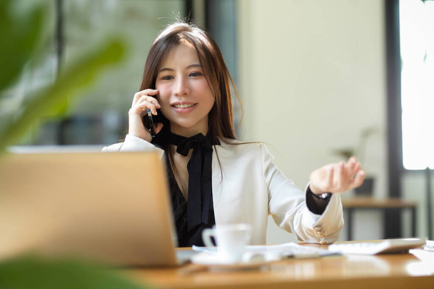 Aziatisch mooi zakenvrouw die zaken doen praten op mobiele telefoon en vragen over iets op de laptop terwijl zitten aan haar bureau. Goed zakelijk gepraat.. - Foto, afbeelding