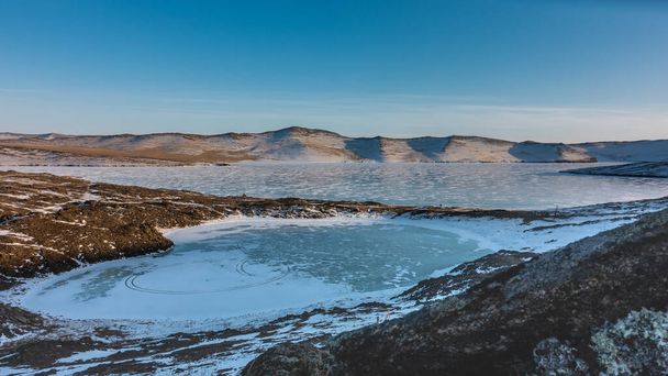 Een geweldig bevroren meer in de vorm van een hart. Op het met sneeuw bedekte ijs zijn cirkelvormige bandensporen zichtbaar. Er is een groot ijsmeer in de buurt. Een bergketen tegen een blauwe lucht. Baikal - Foto, afbeelding