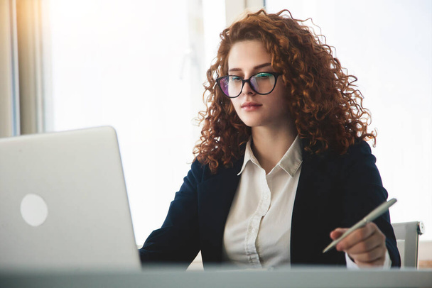 Beautiful young curly red-haired woman in a business suit and glasses works in the office on a laptop - Foto, imagen