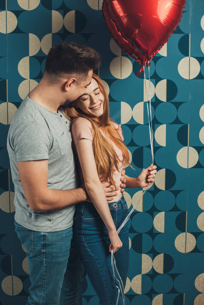 Χαμογελαστό ζευγάρι στέκεται ενώ ποζάρουν με μπαλόνια καρδιά απομονώνονται πάνω από το μπλε φόντο τοίχο. Ημέρα του Αγίου Βαλεντίνου. Όμορφο κορίτσι.. - Φωτογραφία, εικόνα