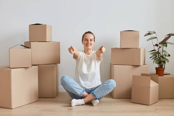 Smiling happy young adult woman wearing white T-shirt sitting on the floor near cardboard boxes with stuff, posing in lotus pose and pointing fingers to camera, choosing you. - Foto, Imagem