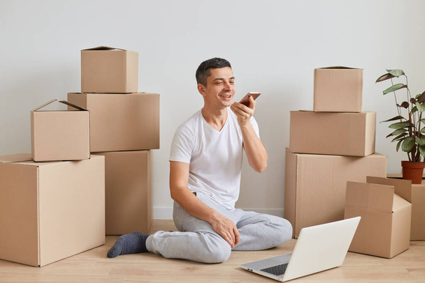 Indoor shot of smiling man wearing white T-shirt sitting on the floor near cardboard boxes with stuff, working on laptop and recording voice messages or commands. - Photo, Image