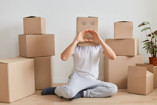 Man wearing white T-shirt sitting on the floor near cardboard boxes with personal belongings, posing with carton box with drawn smiling face and showing heart love gesture with hands, relocation. - Foto, imagen