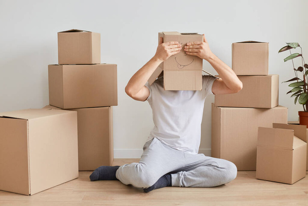 Anonymous unknown man wearing white T-shirt sitting on the floor near cardboard boxes, wears carton box with drawn face on it and covering eyes with palms, having fun during relocating to a new flat. - 写真・画像