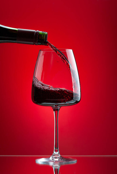 Κόκκινο κρασί ρίχνει από το μπουκάλι στο ποτήρι πάνω από το κόκκινο φόντο - Φωτογραφία, εικόνα
