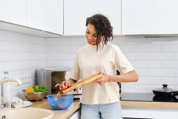 Young african american woman throws chopped tomato into the bowl in the kitchen - Foto, afbeelding