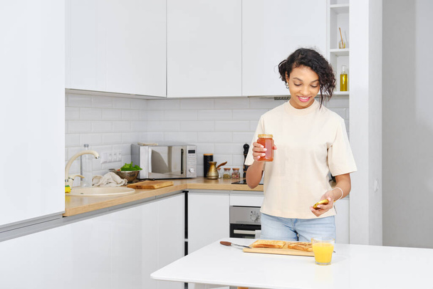Pretty african american woman opens apricot jam to spread it on the toast - Foto, immagini
