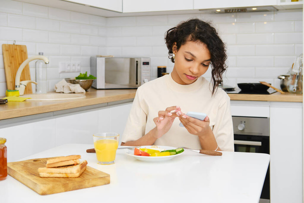 Young african woman reading message on mobile while having breakfast in the morning - Foto, imagen