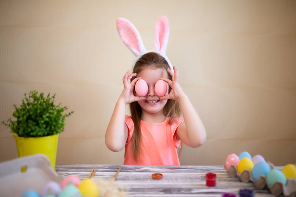 Funny child girl holds easter eggs near the eyes preparing for Easter wearing bunny ears.  - Фото, изображение