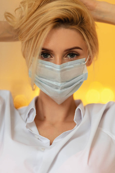 Κοντινό πλάνο πορτρέτο της όμορφης νεαρής ξανθιάς γυναίκας με μια ιατρική μάσκα σε ένα κομψό λευκό πουκάμισο με φόντο τα χρυσά φώτα bokeh. κορίτσι νοσοκόμα εργαζόμενος ματιά στην κάμερα σε εσωτερικούς χώρους - Φωτογραφία, εικόνα
