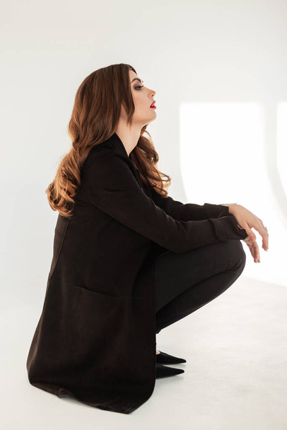 Stilvolle schöne Modell Mädchen mit brünetten Haaren in einem modischen schwarzen Anzug mit Blazer sitzt im Studio auf weißem Hintergrund - Foto, Bild