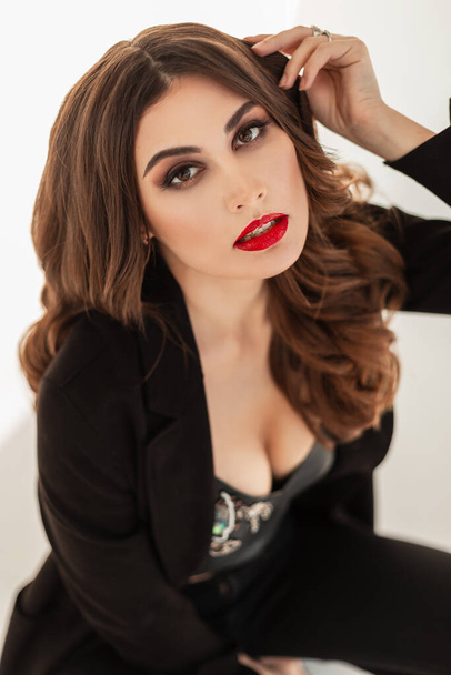 Divatos női portré egy gyönyörű nő göndör haj fényes smink és piros ajkak divatos fekete ruhák a stúdióban nézi a kamerát. Női üzleti stílus. Lány modell - Fotó, kép