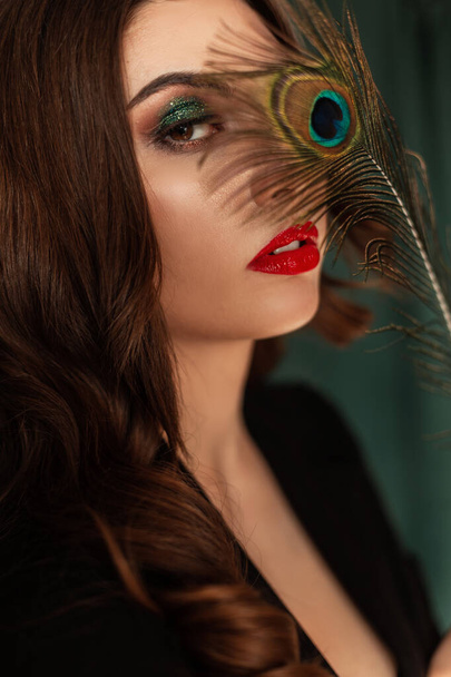 Beautiful young sexy girl model with makeup and hairstyle in a black elegant suit covers her eyes with a colored peacock feather - Valokuva, kuva