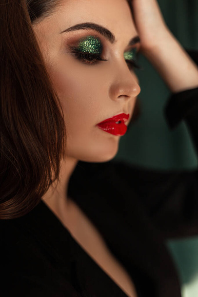 Nahaufnahme Porträt einer hübschen jungen Frau mit professionellem Make-up, roten Lippen und grünen Lidschatten in trendiger schwarzer Kleidung auf grünem Vintage-Hintergrund - Foto, Bild