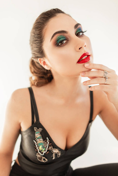 Stylish portrait of a beautiful young sexy woman model with professional makeup, red lips and green shadows in fashionable look with a leather top and breasts sitting on a white background - Fotó, kép
