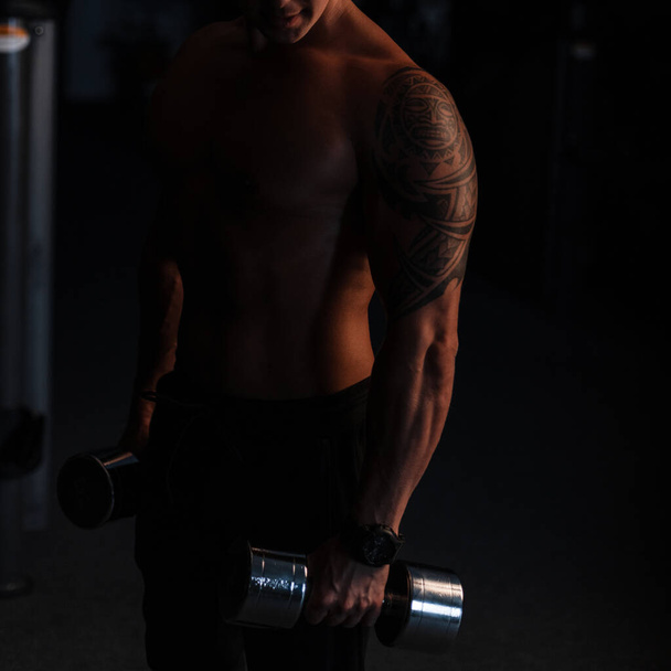 спортивний молодий сексуальний чоловік з голим торсом тримає металеві гантелі в темряві. Концепція спортивного способу життя
 - Фото, зображення