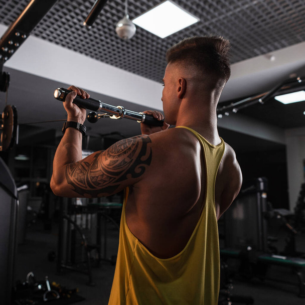 Forte giovane bodybuilder sportivo con tatuaggio in una t-shirt gialla si allena in palestra - Foto, immagini