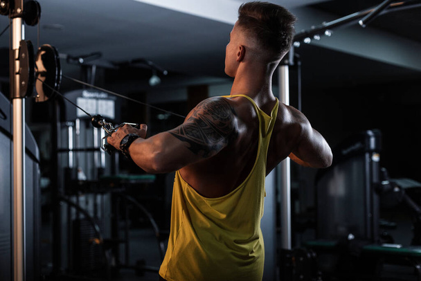 hombre culturista con un tatuaje hace un ejercicio en el gimnasio y bombea su músculo de la espalda en el gimnasio sobre un fondo oscuro. - Foto, imagen