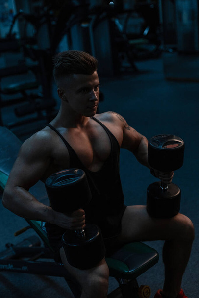 Young athletic handsome man bodybuilder with muscular body in black sportswear sits and does an exercise with dumbbells in the gym on a dark background - Photo, Image