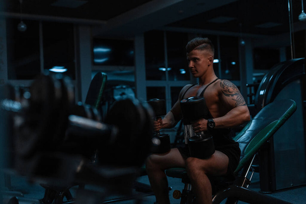 Handsome young man bodybuilder with muscular body sits with dumbbells and pumps muscles in a gym with exercise equipment. Sports lifestyle - Foto, afbeelding