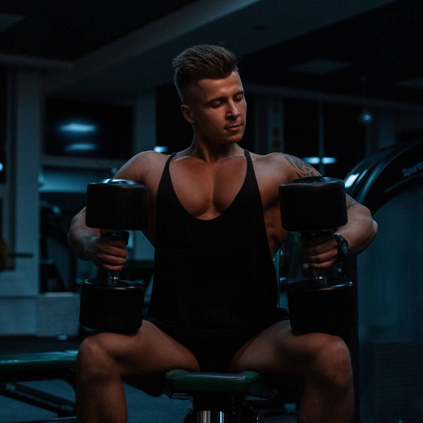 Handsome sporty man bodybuilder with muscular body in fashionable black tank top and shorts sits and does exercises with dumbbells in the gym on a dark background - Photo, Image