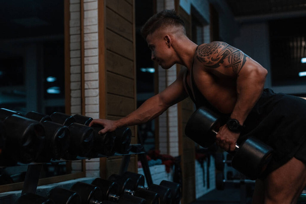 Strong handsome young male bodybuilder with muscular body and tattoo on his shoulder pumping biceps with dumbbells in the gym - Foto, afbeelding