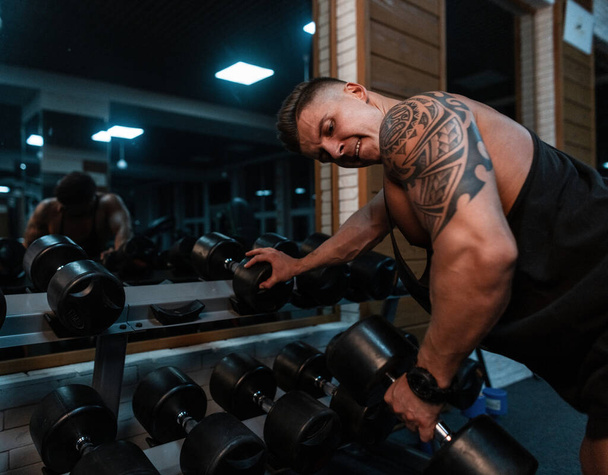 handsome sports man bodybuilder with a muscular body and a maya tattoo pumps up his biceps and exercises with dumbbell in the gym - Photo, image