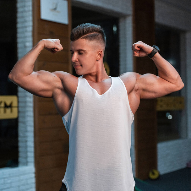 Erőteljes fiatal testépítő férfi izmos test fehér tartály tetején pózol az edzőteremben - Fotó, kép