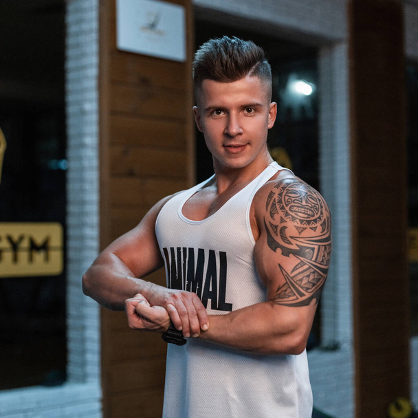 Handsome young man athlete bodybuilder with muscular body and tattoo in white tank top poses in the sport gym - Photo, Image